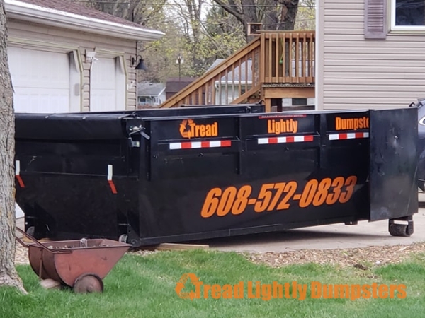 A black dumpster with the words "Tread Lightly Dumpsters" and the phone number "608-572-0833" printed in orange is parked on a driveway. A wheelbarrow sits nearby, and a wooden staircase leads up to a deck in the background, with a house partially visible on the right. The scene is set in a residential area surrounded by trees.