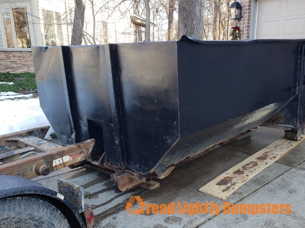 A black dumpster rests on a trailer in a residential driveway. The dumpster is partially obscured by surrounding trees and is positioned on a concrete surface. The side of the dumpster features the text "Tread Lightly Dumpsters" in an orange font. Snow is visible in the background, indicating a cold season.