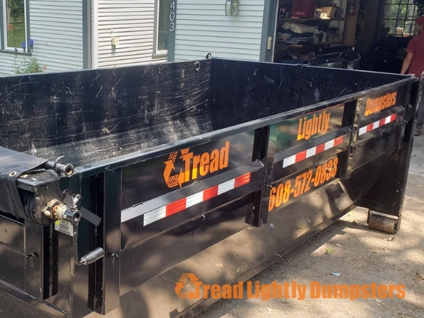 A black dumpster labeled "Tread Lightly Dumpsters" with a phone number, parked outside a house. The background features a light-colored house and a garage filled with various items.