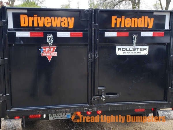 A black dumpster trailer with the words "Driveway Friendly" in large orange letters on the rear doors. There are logos for "FJ Trailers" and "Rollster" on the doors. The bottom of the image features the text "Tread Lightly Dumpsters" in orange. The setting appears to be outdoors, with trees in the background.