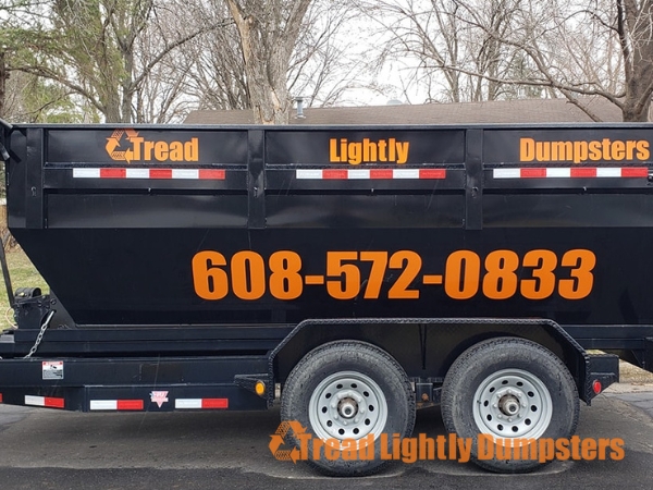 A black dumpster on a trailer, featuring the business name "Tread Lightly Dumpsters" in bold orange letters. The phone number "608-572-0833" is prominently displayed in the center. The background shows a tree and a residential area. The dumpster has reflective strips for visibility and is parked on a paved surface.