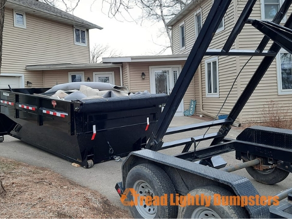 A black dumpster is parked on a driveway in front of a two-story beige house. The dumpster is partially filled with large items, including what appears to be a pile of fabric or padding. Next to the dumpster, a trailer with a stand is visible, used for transporting the dumpster. The house features a porch and large windows, with trees in the background indicating an early spring setting. The text "Read Lightly Bumpsters" is overlayed on the image.