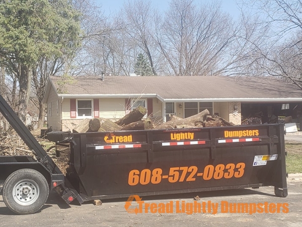 A black dumpster from Tread Lightly Dumpsters is parked on a residential street, filled with debris including logs and branches. In the background, a single-story house with a brown roof and red-trimmed windows is visible, surrounded by bare trees. The dumpster has the phone number 608-572-0833 displayed prominently, along with the company name.