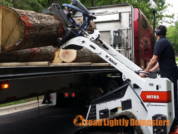 A Bobcat MT85 compact skid-steer loader is lifting large tree logs onto a truck. The operator, wearing headphones, is focused on maneuvering the equipment. The truck is equipped with a dump bed, and the setting appears to be a residential area with greenery in the background. The logo “Tread Lightly Dumpsters” is visible in the corner.