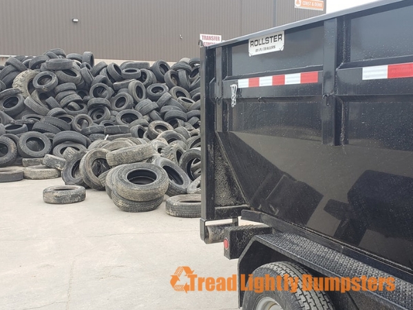 A large pile of used tires is stacked in the background, while the foreground features a black dumpster labeled "ROLLSTER" with a logo for "Tread Lightly Dumpsters." The scene is set in a parking lot or transfer station, showcasing the process of tire disposal and recycling.