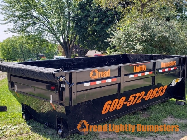 A large black dumpster with orange text reading "Tread Lightly Dumpsters" and a phone number (608-572-0833) is parked on grass, surrounded by trees and bushes. The dumpster has a tarp cover and visible wheels.