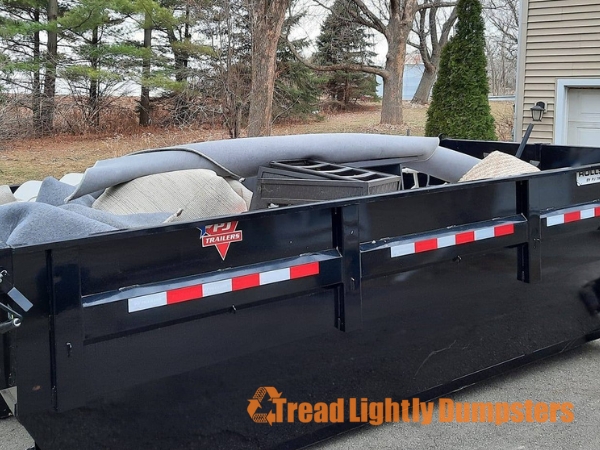 A filled black dumpster sits in an outdoor area, containing various items such as furniture and a rolled-up carpet. In the background, trees are visible, and there is a house with a light-colored exterior. The logo for "Tread Lightly Dumpsters" is displayed prominently at the bottom of the image.