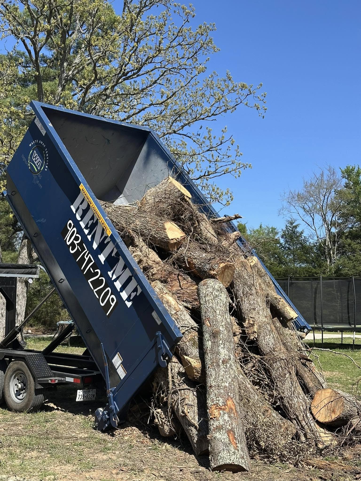 Blue roll-off dumpster from Waste Away Service unloading a large pile of tree logs and branches on a clear day.