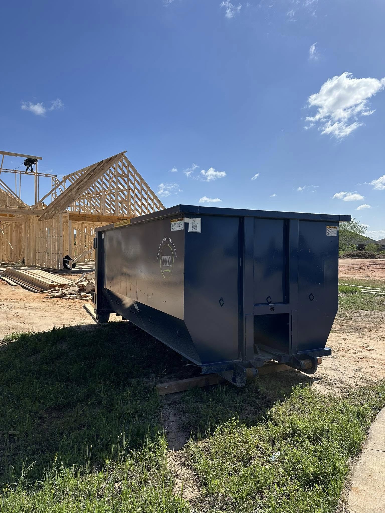 Blue roll-off dumpster from Waste Away Service placed at a residential construction site with visible wooden house framing.
