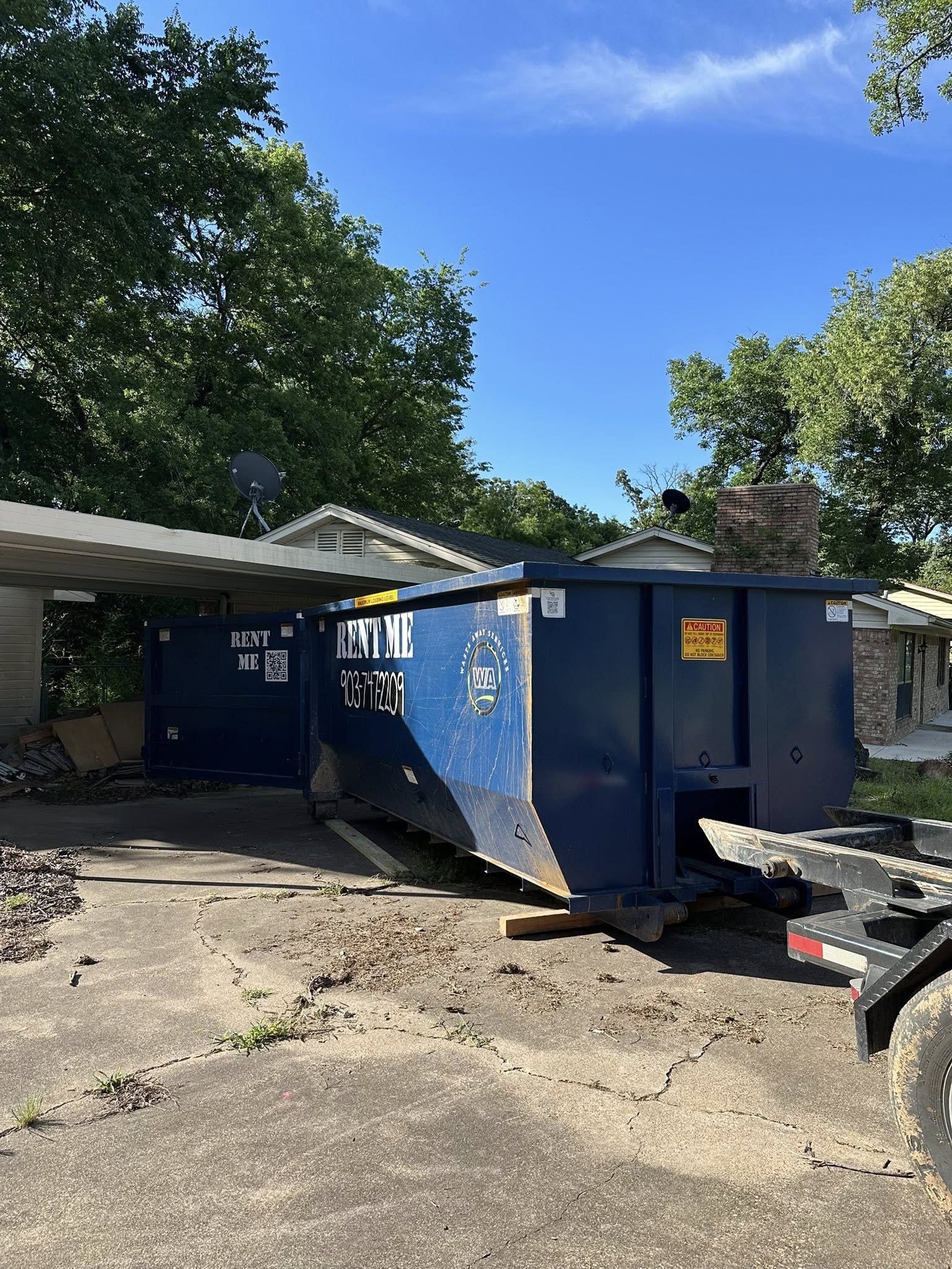 Blue dumpster with "RENT ME" and phone number branding placed in a residential driveway under a clear sky.